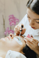 Girl cosmetologist laminates and paints woman eyelashes