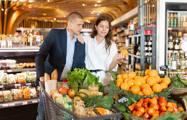 Glad couple is standing with cart with products in the supermarket