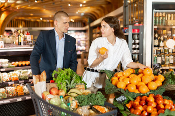 Glad couple is standing with cart with products in the supermarket