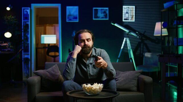 Bored man relaxing at home on couch, munching on popcorn while passing time watching movies on TV. Lazy person eating snacks and zapping through TV channels in RGB lit living room, camera B