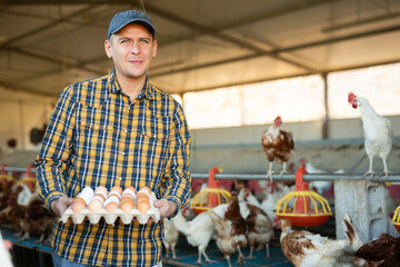 Happy successful owner of poultry farm engaged in breeding of laying hens standing in chicken coop, holding carton tray of fresh organic eggs