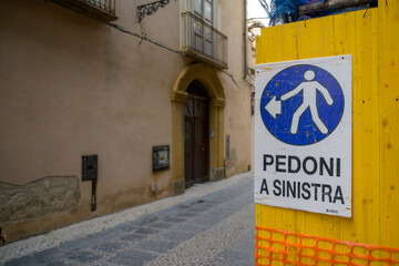 Italian Pedoni a Sinistra, Pedestrians to the left blue and white sign on wooden yellow background in village street, Cefalu, Sicily.