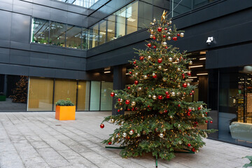 Beautifully decorated Christmas tree with red and gold ornaments standing in front of sleek modern office building, creating a cozy holiday atmosphere in an urban business district.