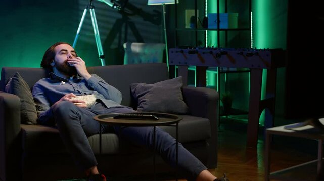 Bored man at home entertaining himself by throwing popcorn in air and catching it with mouth, wasting time. Person slacking off, eating salty snacks and lounging on couch watching TV, camera A