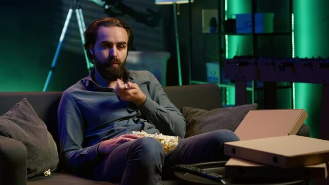 Exhausted man watching shows on TV late at night begore going to sleep and eating popcorn. Sleepy man in pajamas eating snack on couch, consuming mindless entertainment, camera A