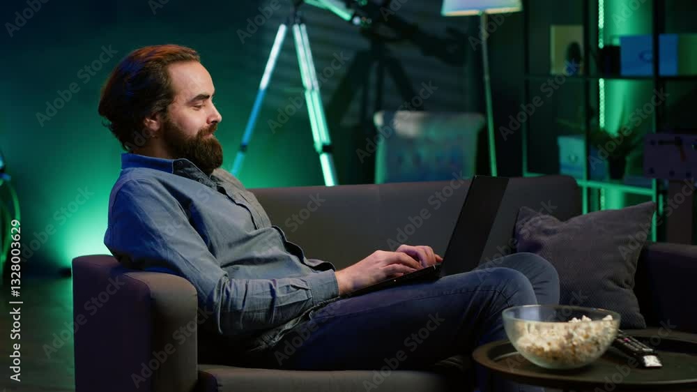 Aufkleber man working on laptop lounged on neon lit apartment couch while picking tv channel to watch as distr