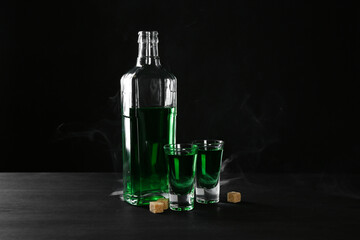 Absinthe in shot glasses, bottle and brown sugar on table against black background with smoke