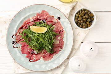 Delicious beef carpaccio with arugula, cheese, lemon and capers on white wooden table, flat lay