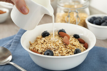 Woman pouring milk into bowl with oatmeal, blueberries and almonds at table, closeup