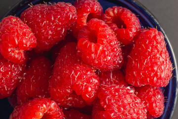 A high-resolution macro shot of ripe raspberries in a blue glass bowl, covered in water droplets,...