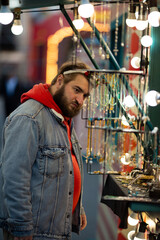 Vertical city photo. Young adult white man with beard, carnival deer horn on Merry Christmas fair near street market stall with jewelry. Concept of winter holiday gift, sale, choice, Happy New Year