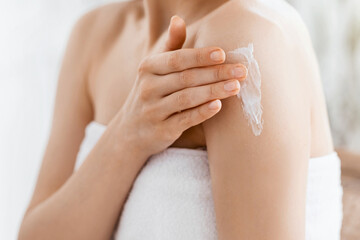 Closeup of young woman wrapped in towel applying body lotion on shoulder after shower, copy space