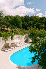 White sun loungers stand on the shore of an oval pool in a green garden