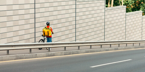 Takeaway food and delivery in city. Courier with big yellow backpack walking with bicycle along track in city, back, copy space
