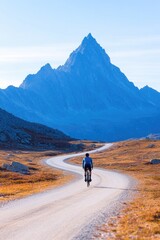 Obraz premium lone cyclist rides winding mountain road surrounded by towering peaks under clear blue sky