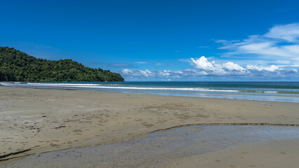 A beautiful beach on a tropical island. The stream flows to the sea. There are sand bubbler crabs balls on the sand. The waves of the turquoise ocean are foaming. A hill against the blue sky, clouds. 