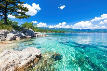 Fototapeta premium Clear turquoise water laps gently against rocky shoreline, under a vibrant blue sky dotted with fluffy white clouds. Mountains in distance