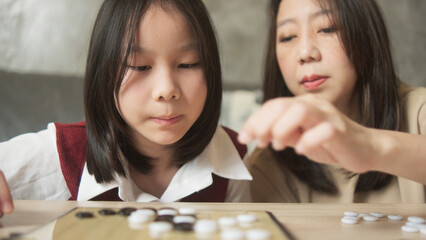 Fototapeta premium Close-up shot, Asian family times, mom, and kid fun playing Go, checkers board game on table in home living room together with love and cheerful, happy weekend relaxing hobby of child and parent.