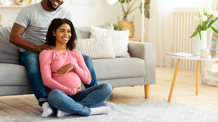 Obraz premium Pregnant black woman feeling relaxed, sitting on floor at living room, attentive husband massaging her shoulders, free space