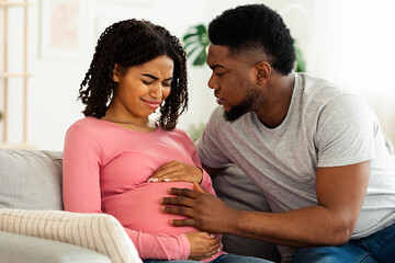 Fototapeta premium Attentive african husband supporting his pregnant wife in pain, home interior