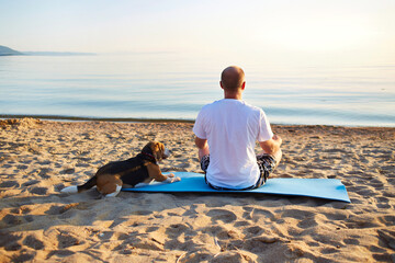 A man sits on a sandy beach on the seashore in the lotus position. A beagle dog is nearby....