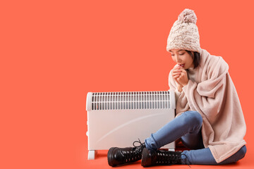Young Asian woman in winter clothes with radiator sitting on orange background