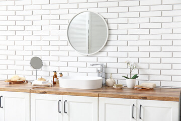 Sink, bottles of cosmetic products on chest of drawers and mirror on white brick wall in bathroom