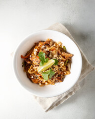 Traditional Pad Thai with stir-fried rice noodles, tender chicken, vegetables, and peanuts, garnished with fresh cilantro and a lime wedge. Served in a white bowl on a white table, close-up view. High