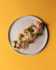 On a bright yellow background, a white plate of pasta featuring tender salmon, fresh broccoli, and black olives. A vibrant seafood dish full of color and flavor