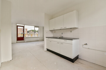 Spacious and minimalistic kitchen design with white cabinets and gray countertops, featuring unfinished flooring and large windows for natural light.