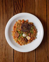 Traditional Uzbek plov pilaf with fragrant rice, tender beef, carrots, and spices, garnished with green onions, quail egg, and garlic. Served on a white plate over a rustic wooden table, top-down view