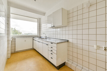 A bright and simple kitchen featuring white tiles, functional cabinets, and a serene view through the window, embodying minimalist design principles.