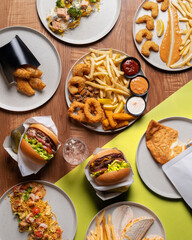 Top-down view of multiple fast-food dishes, including burgers, fries, fish and chips, and various condiments, arranged on a wooden table.