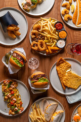 Top-down view of multiple fast-food dishes, including burgers, fries, fish and chips, and various condiments, arranged on a wooden table.