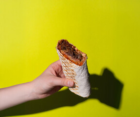 A horizontal photo of a hand holding a stuffed shawarma with grilled meat, crunchy vegetables, and tangy sauce, set against a vibrant green background. Ideal for marketing or commercial purposes.