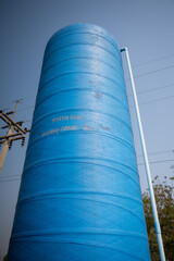 tall blue water tank stands against clear sky, with pipe attached to its side, near power lines and trees