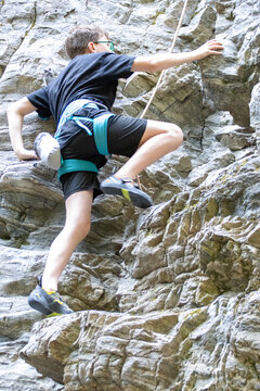 Young boy rock climbing with safety gear on a rugged outdoor cliff.