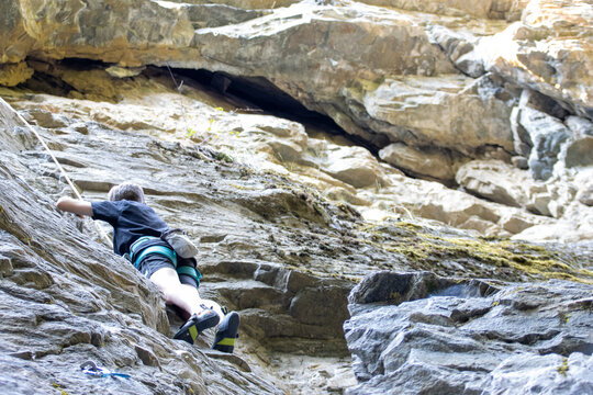 Young boy rock climbing with safety gear on a rugged outdoor cliff.