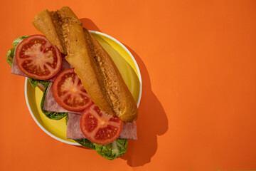 Top view of a sandwich with bread, lettuce, ham, and tomato on a yellow plate against an orange background. Sandwich on a colorful background