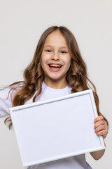 Fototapeta premium White empty banner. Happy smiling little school girl holds white blank paper. Young smiling woman show blank board. Close up female model portrait on white background. Mock up. Copy space.