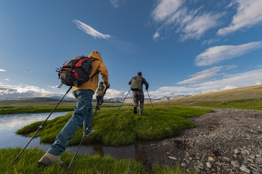 Tourists and travelers run through a green meadow next to a river against a backdrop of snowy mountains, carrying backpacks on their shoulders.