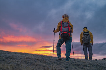 Travelers in bright clothes with backpacks on their backs and trekking poles in their hands stand on the mountain during the fiery sunset, which forms in the golden hour. A golden hour with people on 