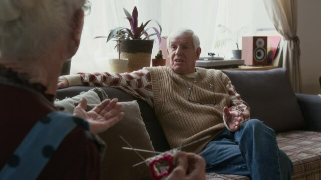 Rack focus shot of elderly woman knitting with needles and talking with senior male friend relaxing on sofa in cozy living room during day in nursing home