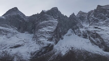 Sideways drone shot flying underneath Troll Wall or Trollveggen in Norway with trees in the forground looking at the snowy mountain tips on a bright day near sunrise LOG
