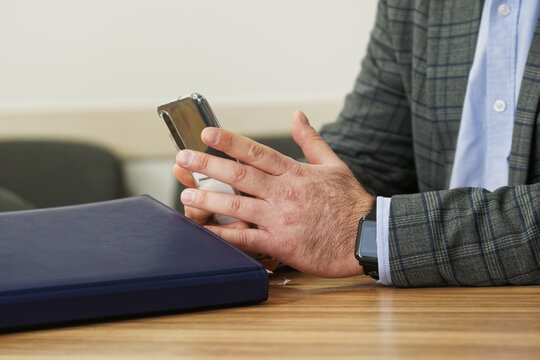 Close-up of a man in a suit jacket using a smartphone next to a folder on a desk, symbolizing modern business negotiations in an office environment. Photo