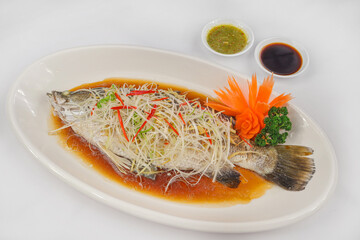 A whole steamed fish served with soy-based sauce, garnished with shredded ginger, chili, and fresh herbs, accompanied by two dipping sauces and carved vegetables.