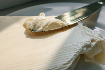 Close-up of a knife spreading fresh butter with soft curls forming on the blade. Warm natural light highlights the creamy texture, creating a rustic and appetizing food scene