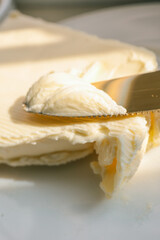 Close-up of a knife spreading fresh butter with soft curls forming on the blade. Warm natural light highlights the creamy texture, creating a rustic and appetizing food scene