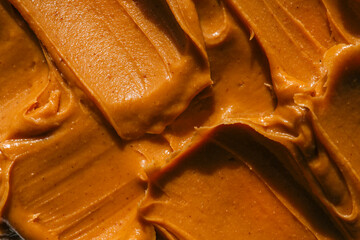 Close-up of creamy peanut butter spread with a rich, smooth texture. The golden-brown surface has visible swirls and ridges, showcasing its thick and glossy consistency