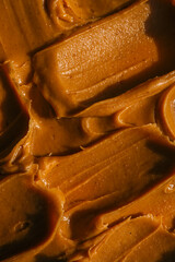 Close-up of creamy peanut butter spread with a rich, smooth texture. The golden-brown surface has visible swirls and ridges, showcasing its thick and glossy consistency